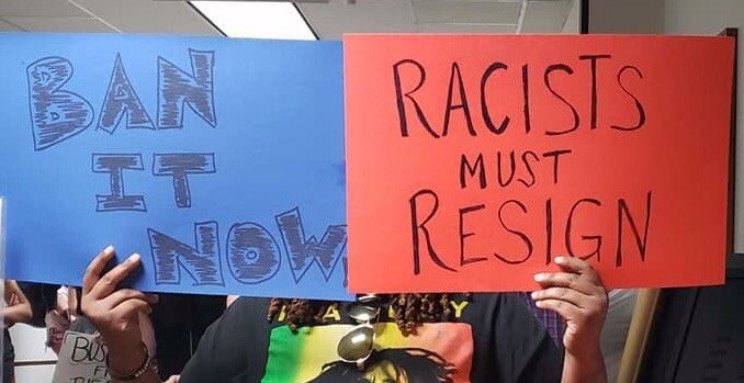 PHOTO: Two signs held by a black person inside a building. The blue rectangular poster on the left read in black, all-caps letters, "BAN IT NOW!" The orange, rectangular sign on the right reads in black, all-caps letters, "RACISTS MUST RESIGN". The person holding the signs is wearing a black t-shirt with a photo of Bob Marly over a green, yellow, and red background. The person's face is hidden behind the signs. The legs of other people with signs can be seen in the background.